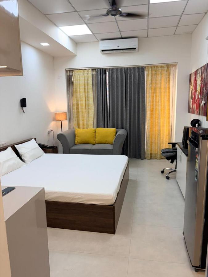 Deluxe studio with a double bed, grey sofa with yellow cushions, wall AC, work chair and a fridge by the wall